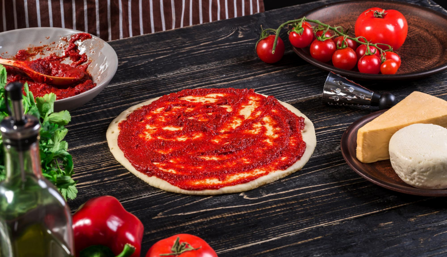 Připravená pizzová základna s rajčatovou omáčkou a čerstvými surovinami.