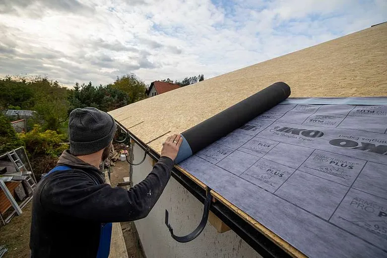 Muž natáčí hydroizolační fólii na šikmou střechu z OSB desek.