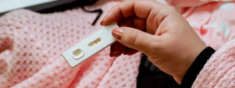 Ruce drží těhotenský test s pozitivním výsledkem na růžovém pozadí.