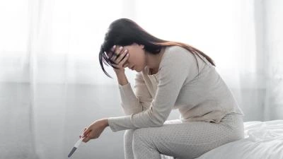 Žena sedí na posteli s pozitivním těhotenským testem v ruce.