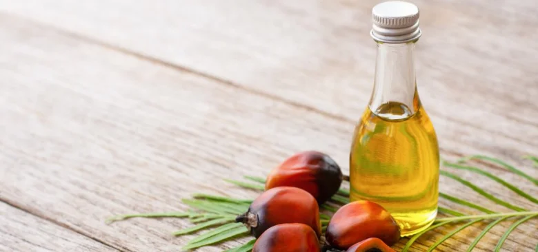 Sklenička s palmovým olejem vedle čerstvých plodů palmy na dřevě.