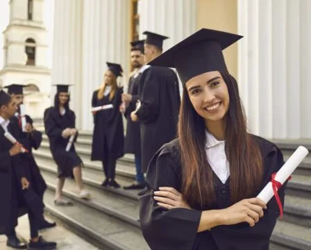 Usměvavá žena v šatě s diplomem na schodech před univerzitou.