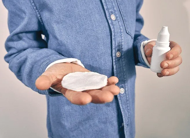 Osoba drží kapky do očí a vatový tampon pro léčbu zánětu spojivek.