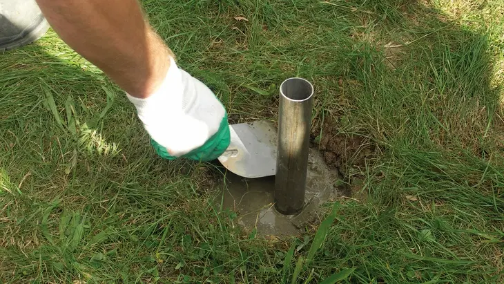 Člověk míchá suchý beton kolem kovové trubky na trávníku.