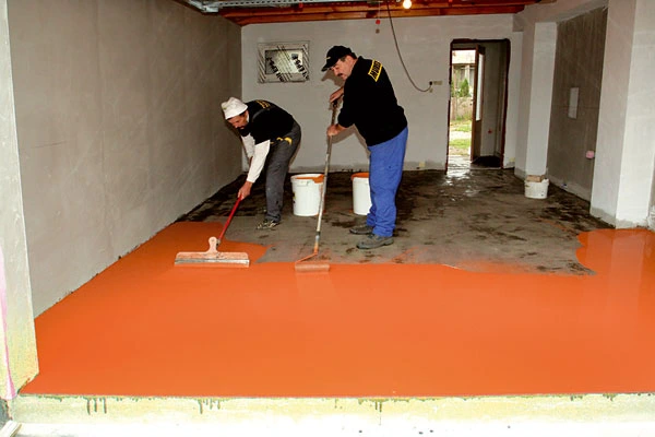 Dva dělníci nanášejí oranžovou epoxidovou podlahu v nezařízené místnosti.